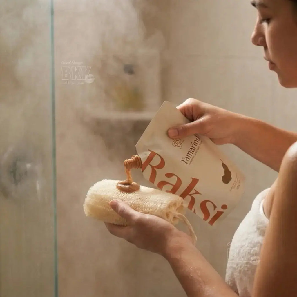 Person applying soap to a sponge with 'Raksi' branding in a bathroom setting.